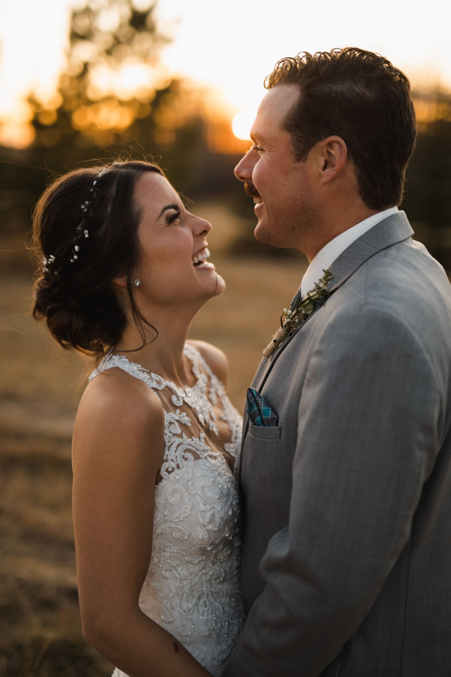 Sunset on wedding day, bride is wearing lace dress and laughs as she gazes at her husband lovingly.