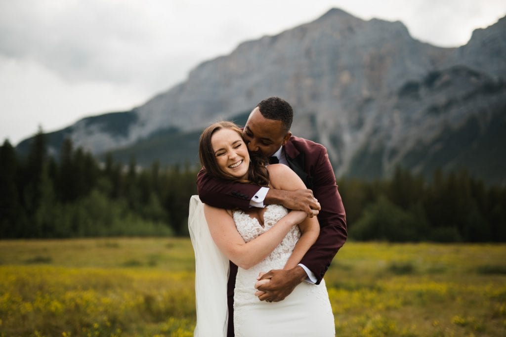 Groom snuggles up to Bride in a big bear hug! Romantic photos in front of beautiful Banff mountains, for their summer covid elopement.