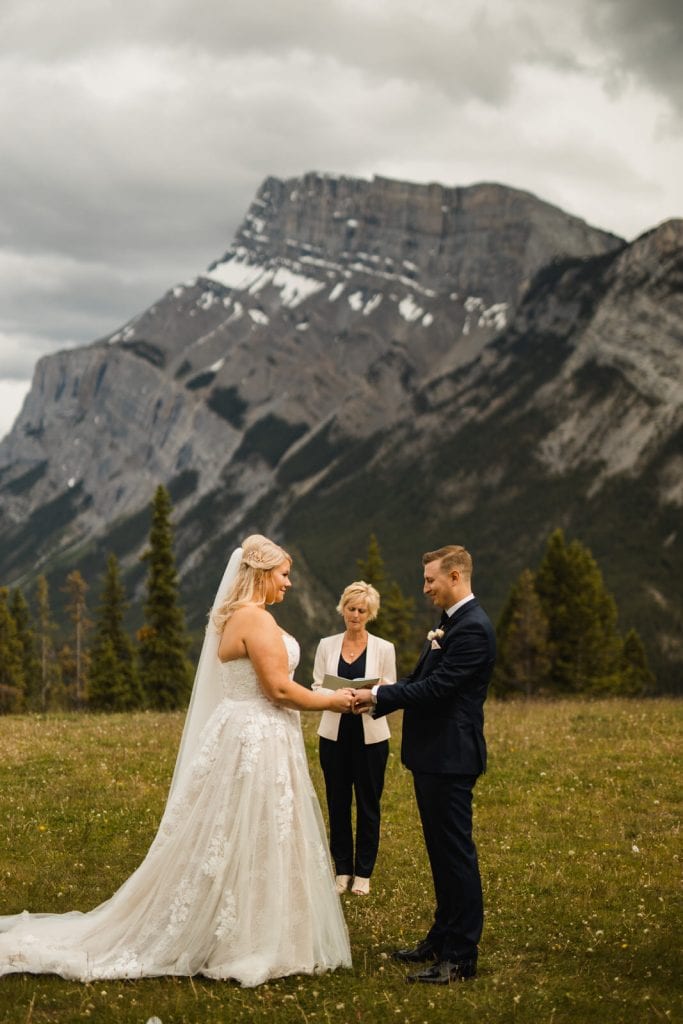 Couple elopes at tunnel mountain in Banff, Alberta. Their beautiful summer wedding was softly lit with the clouds, it made the mountain Rundle look absolutely stunning. Bride and Groom share vows with their officiant, at a gorgeous Banff location.