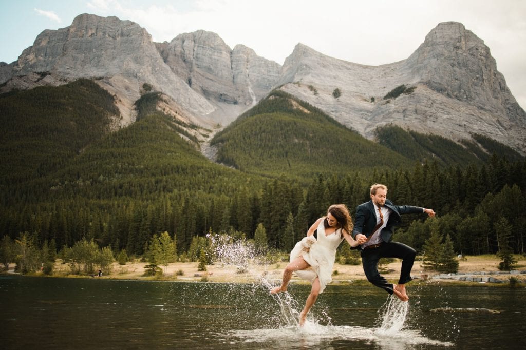 Couple jumps for joy during trash the dress session in Banff mountains. Mountain fed lake is extremely cold, but bride and groom run into the lake anyways