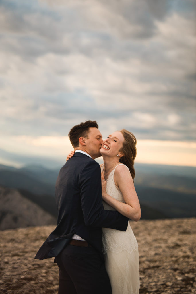 Bride and groom eloping on the top of a mountain, her hair is blown by the wind, she is wearing a white lace dress and he is wearing a blue suit