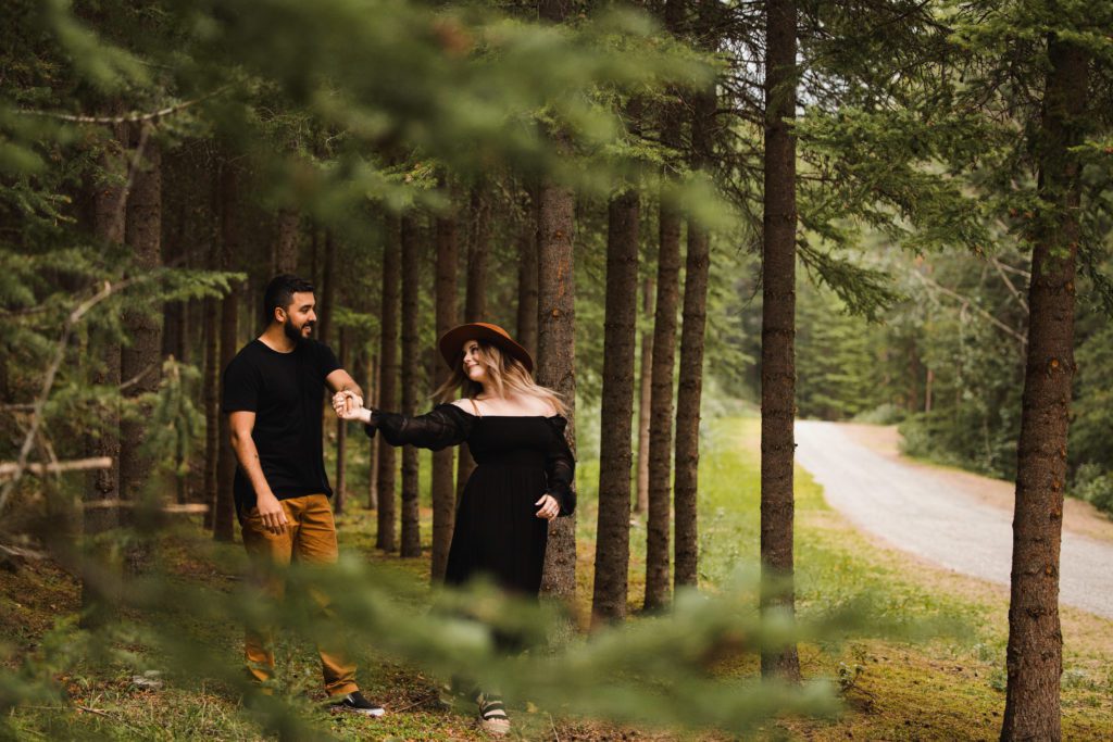 couple wander through a forrest of pine trees for their engagement photos. Her outfit is a cute black dress with a tan hat, while he is wearing a black t-shirt and tan pants that match her trendy hat