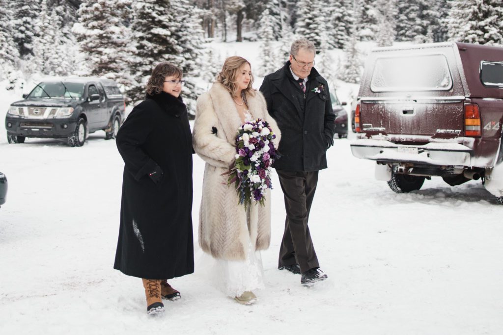 Bride walking up the aisle with her parents in the deep snow for her outdoor winter ceremony