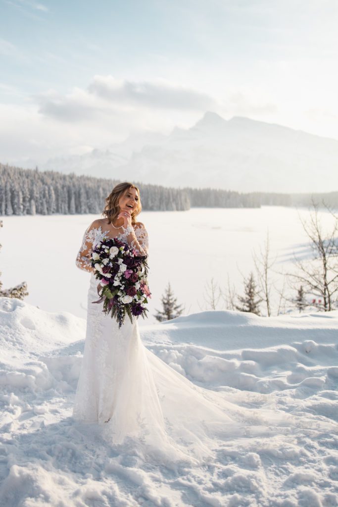 Stunning bridal portrait, with a gorgeous lace wedding dress, a fresh layer of deep snow on the mountain lake, and jagged mountains in the background