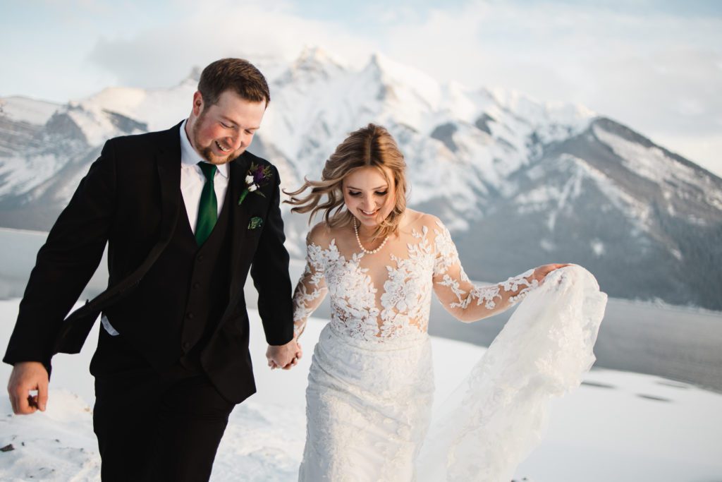 couple walking and holding hands with stunning mountain backdrop on their magical winter wedding day in banff