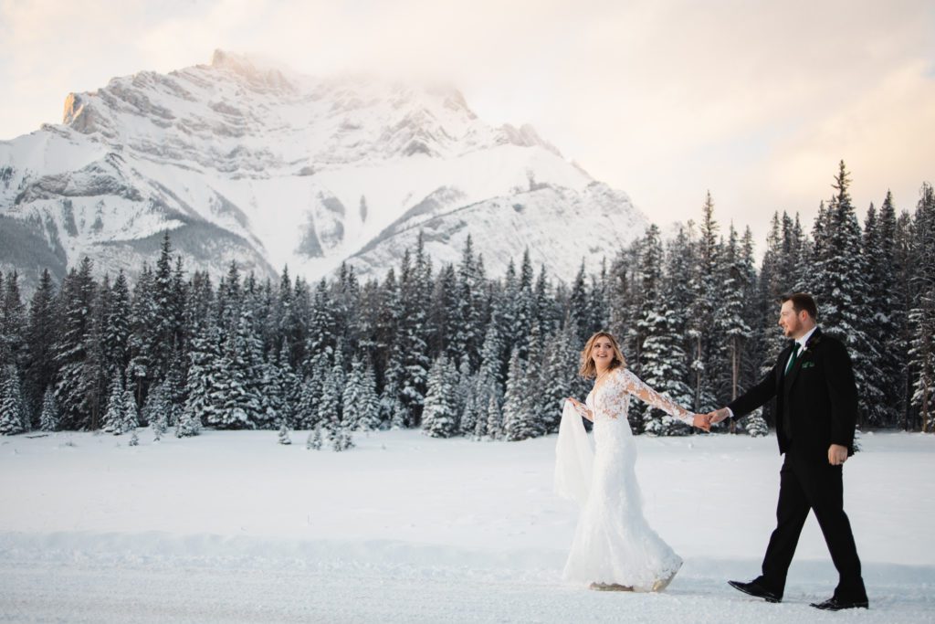 winter wedding in Banff national park, mountains covered in snow, couple walk across a snowy valley with her long sleeve lace dress