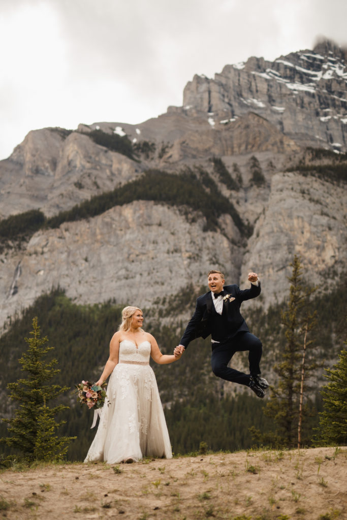 Groom jumps for joy after marrying bride. Mountain backdrop, she is wearing a stunning essence of australia lace wedding dress. Documented by your Banff wedding photographer