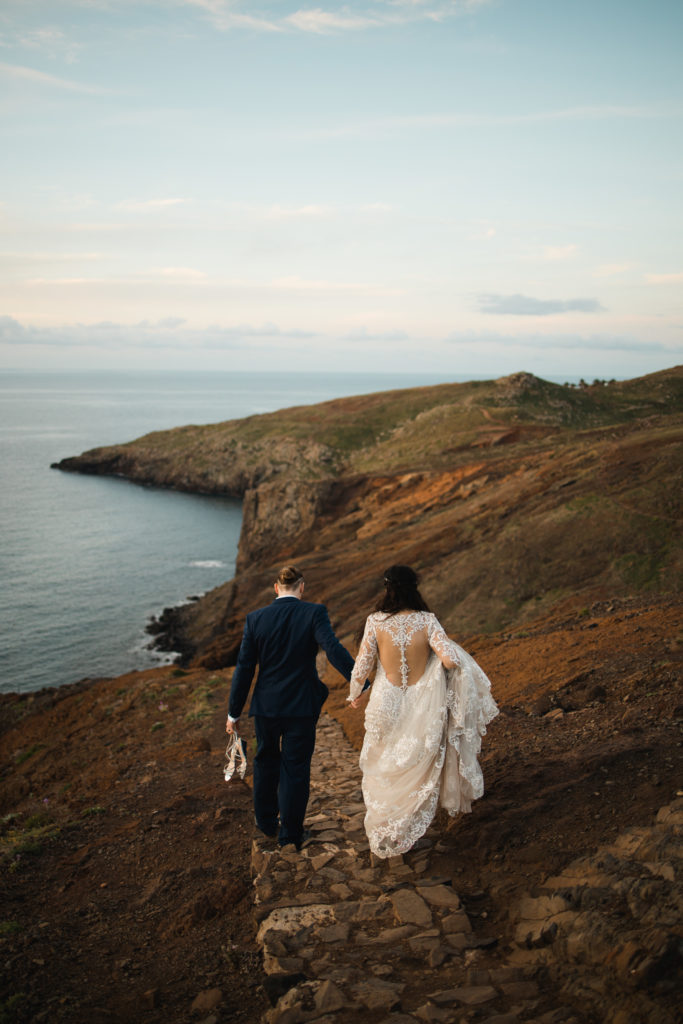 bride and groom near the ocean walking away while holding hands. groom is holding the brides heels while she holds her long lace wedding dress