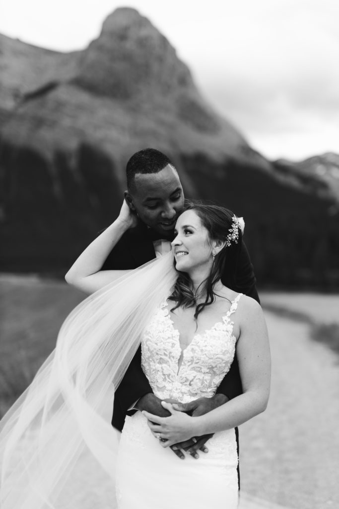black and white photo of bride and groom in the mountains, the wind is blowing her veil across her lace wedding dress. Photographed by your Banff wedding photographer.